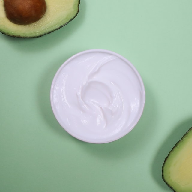 A top-down view of a white bowl filled with creamy, white avocado body butter, surrounded by fresh, halved green avocados on a pale green background.