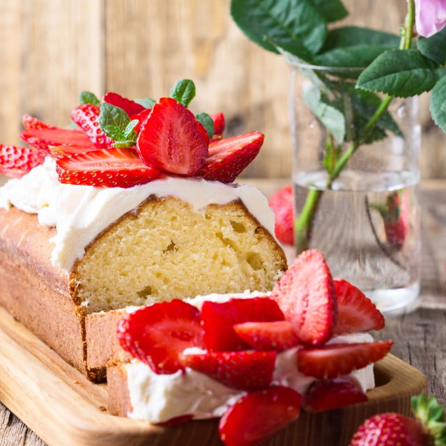 A thick slice of golden pound cake topped with a generous dollop of whipped cream and sliced fresh strawberries, set against a clean, light background.