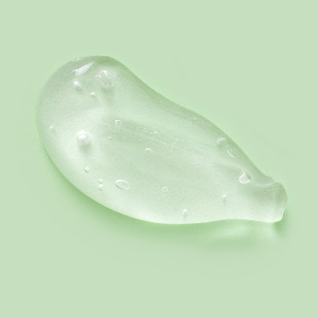 A thick, clear, glossy swatch of transparent shampoo gel with small suspended air bubbles, resting on a solid pale green background.