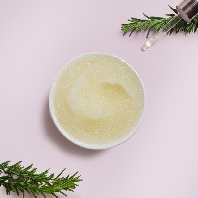 A top-down view of a white bowl filled with granular, pale yellow sugar scrub base, framed by fresh green rosemary sprigs and a glass dropper on a pastel pink background.