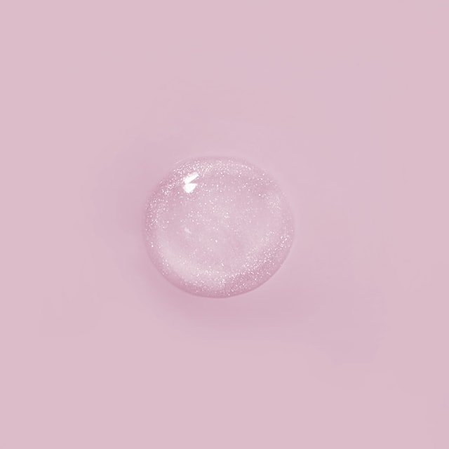 A top-down close-up of a perfectly round, clear puddle of liquid packed with fine, sparkling silver shimmer on a solid pastel pink surface.