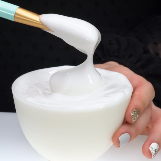 A close-up of a person holding a white bowl while a makeup brush lifts a thick, glossy dollop of white facial cream, demonstrating its rich elasticity.