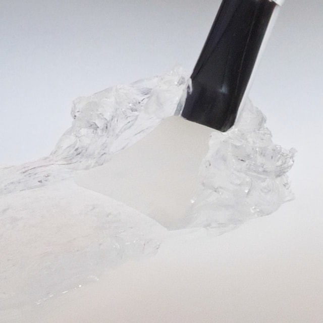 A close-up of a black-handled tool lifting a thick, highly structured dollop of crystal-clear hair pomade, showcasing its firm, jelly-like texture against a light background.