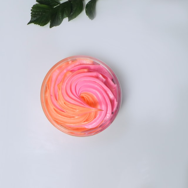 A top-down view of a clear cosmetic jar filled with bright neon pink and bright orange swirled body souffle, set against a clean white background with a single green leaf.