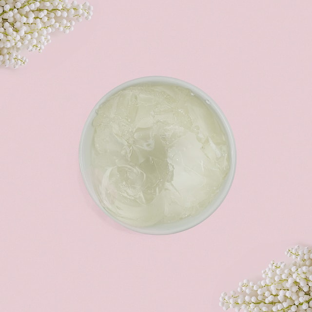 A top-down view of a white bowl filled with chunky, transparent jelly soap base, framed by delicate white flowers in the corners against a pastel pink background.