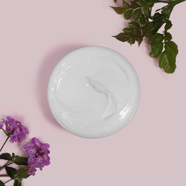 A top-down view of a white bowl filled with smooth, white massage lotion, gracefully framed by green leaves and delicate purple flowers on a pastel pink background.