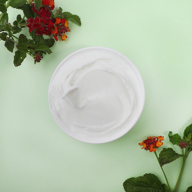 A top-down view of a white bowl filled with a smooth, thick white body custard base, framed by vibrant red and orange flowers and green leaves on a pastel green background.
