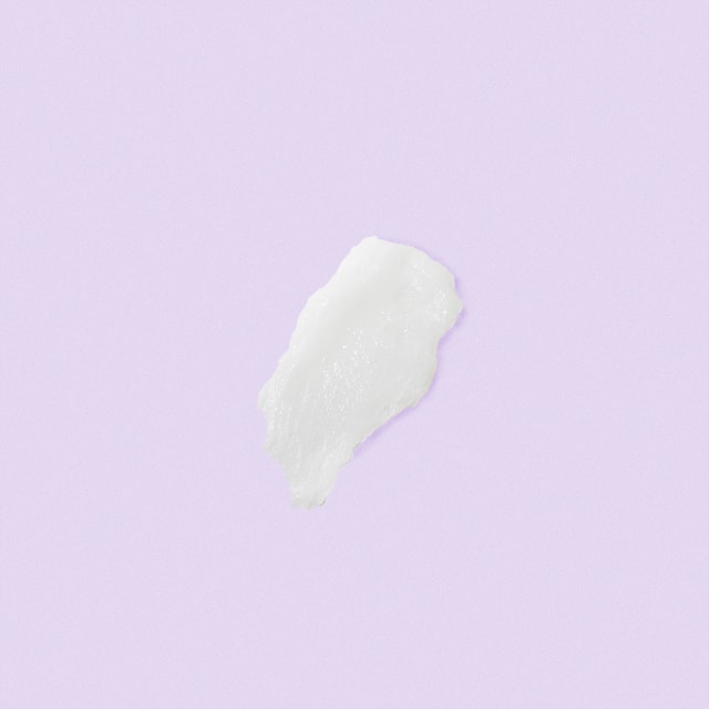 A top-down close-up of a smooth, thick swatch of white hair conditioner resting on a solid pastel purple surface.