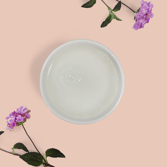 A top-down view of a white bowl filled with a clear, lightweight dry oil base, gracefully framed by delicate purple flowers and green leaves on a soft peach background.