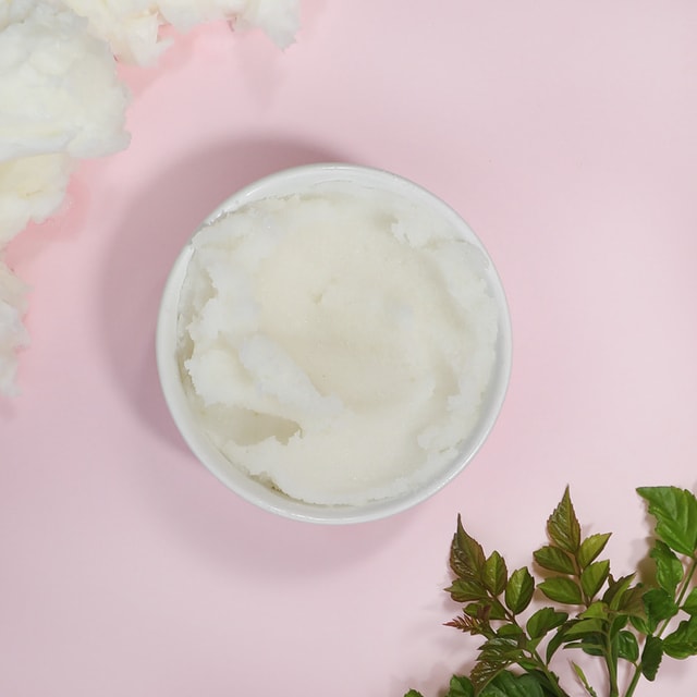 A top-down view of a white bowl filled with a bright white granular body polish, gracefully framed by raw shea butter chunks and fresh green leaves on a pastel pink background.