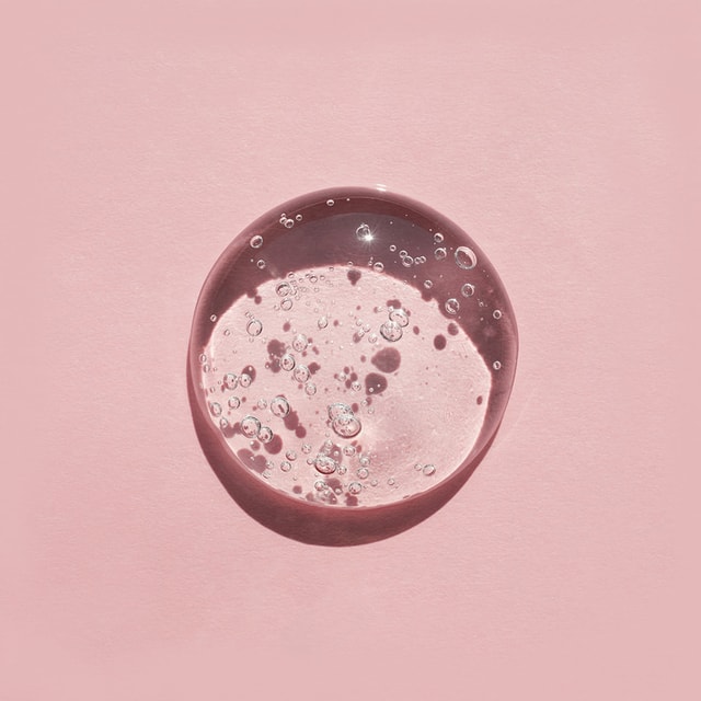 A top-down close-up of a perfectly clear, thick swatch of suspension shower gel packed with suspended air bubbles, casting a distinct shadow on a solid pink surface.