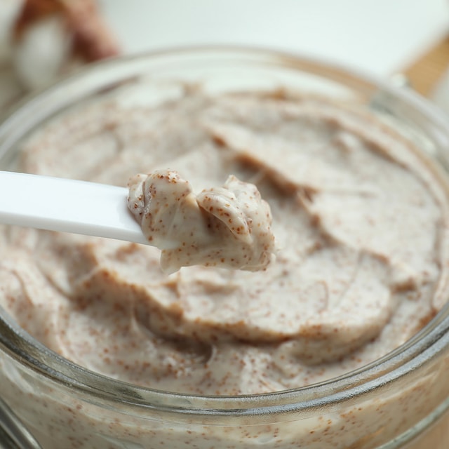 A close-up of a white cosmetic spatula lifting a thick, creamy dollop of walnut body scrub out of a clear glass jar, highlighting its dense, moisturizing texture.