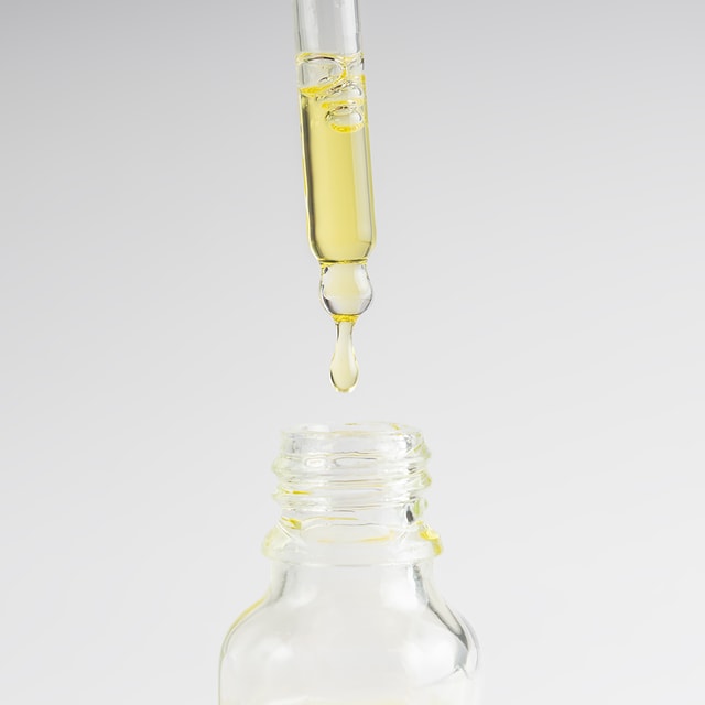 A close-up of a glass dropper dispensing a single drop of clear, pale yellow dry oil into the neck of a clear glass bottle against a clean white background.
