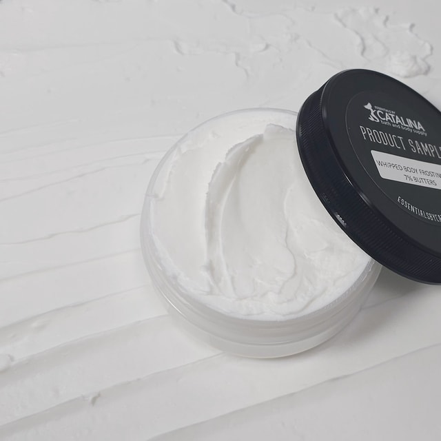 A top-down view of a clear cosmetic jar filled with thick, swirled white body cream with its black lid resting nearby, sitting on a surface completely covered in a textured smear of the same white cream.