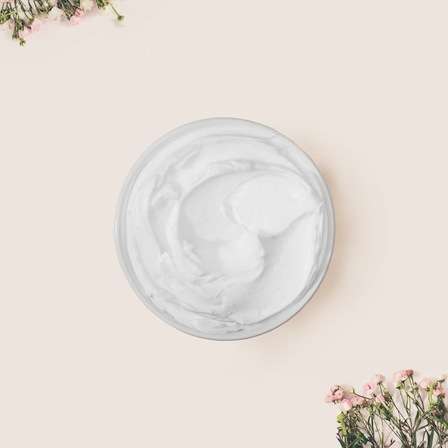 A top-down view of a white bowl filled with a thick, fluffy, bright white whipped soap base, framed by delicate pink flowers in the corners against a soft beige background.