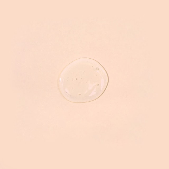 A top-down close-up of a perfectly round, clear puddle of liquid wash base featuring tiny suspended bubbles, resting on a solid pastel peach surface.