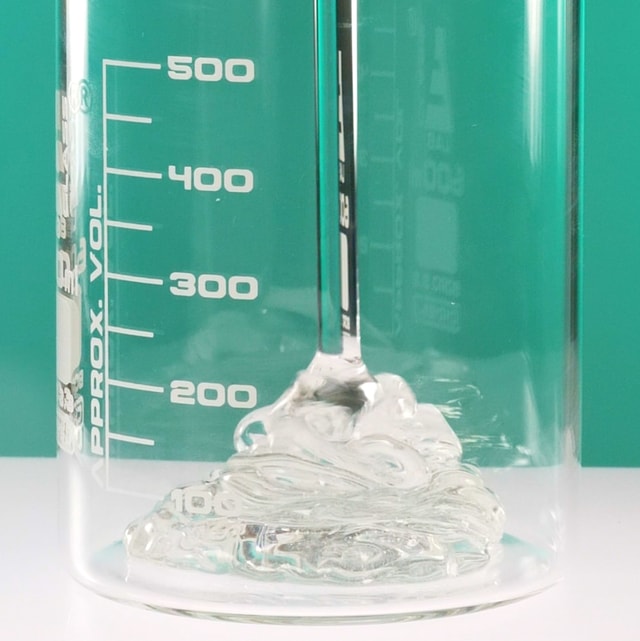 A close-up of a clear, thick liquid face wash gel pouring and pooling into a clear glass measuring beaker with white volume markings against a solid teal green background.